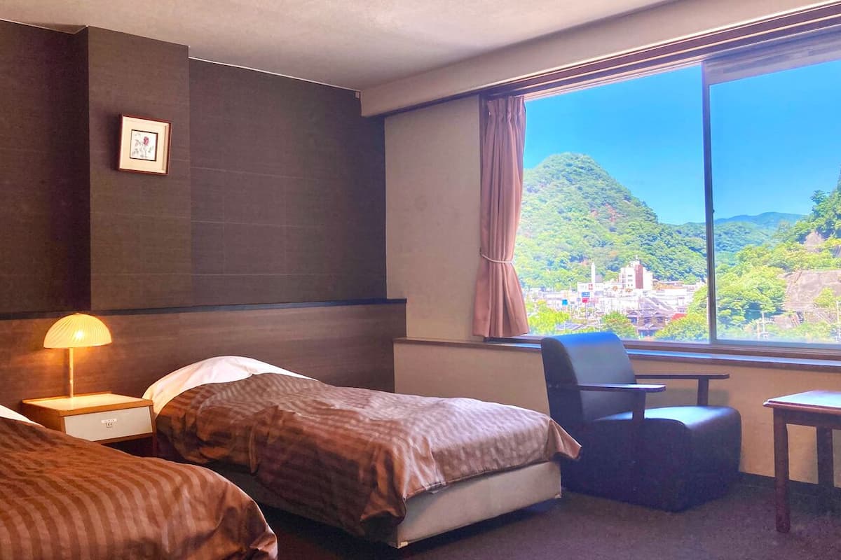 Habitación de hotel con dos camas, un escritorio, una silla y un ventanal con vistas a un paisaje montañoso.