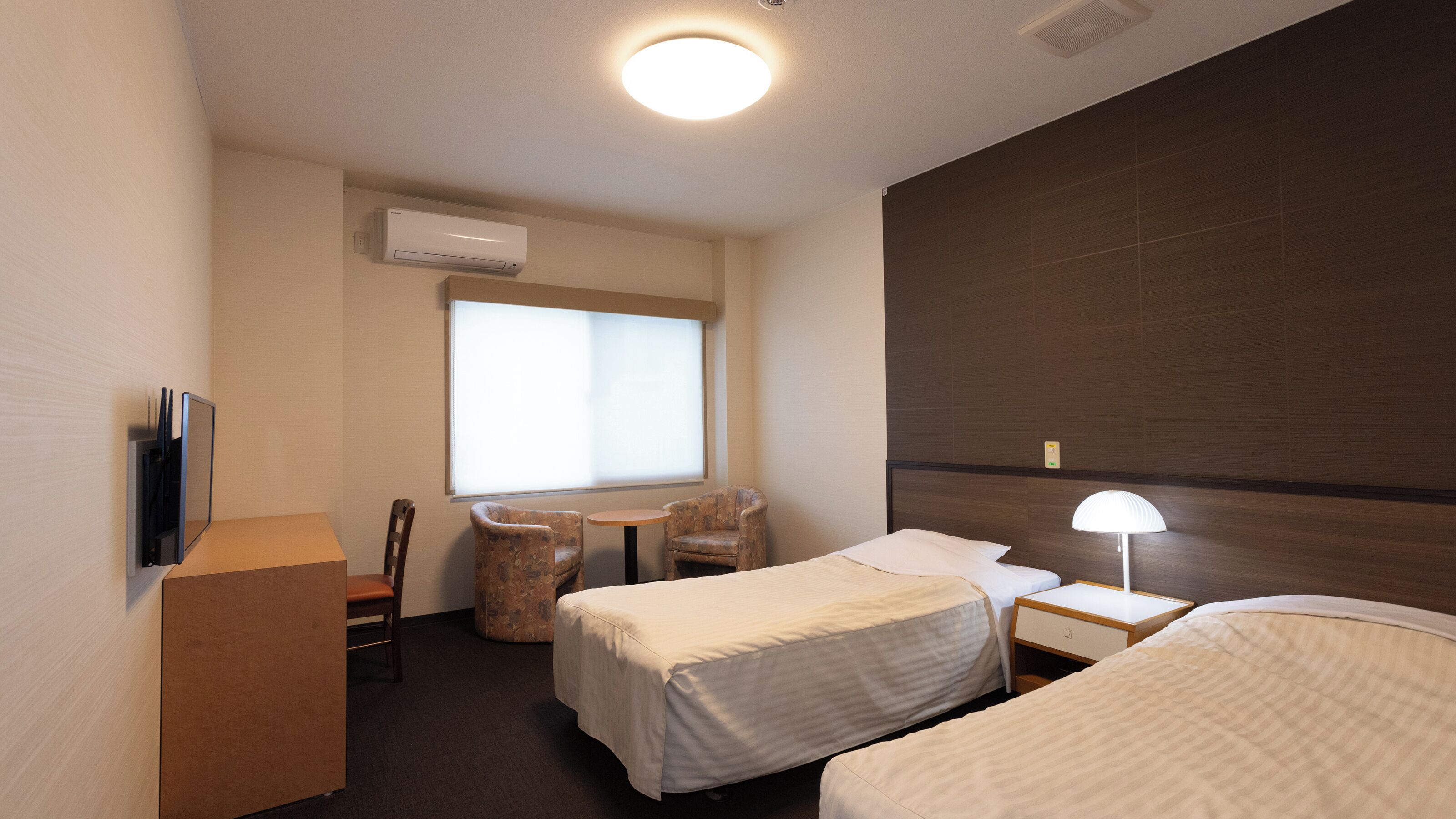 Ein Hotelzimmer mit zwei Betten, einem Schreibtisch, einem Stuhl, einem Fernseher und einer Klimaanlage.