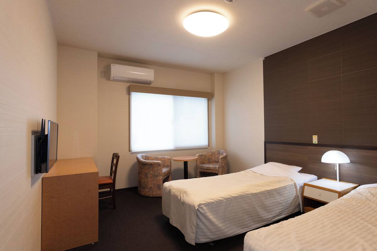 Quarto de hotel com duas camas, uma mesa, uma cadeira, uma televisão e aparelho de ar condicionado.