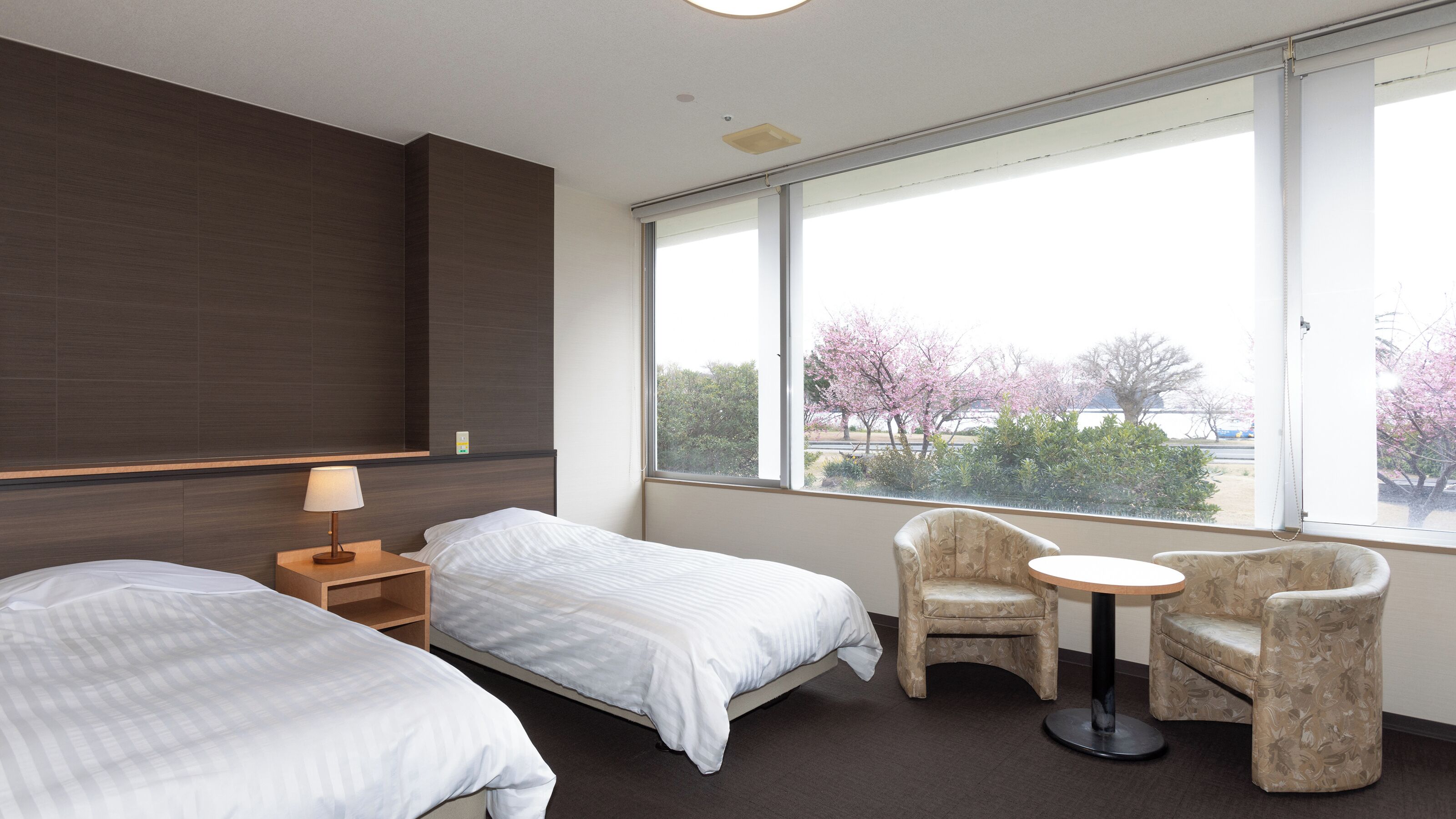 Ein Hotelzimmer mit zwei Betten, einem kleinen Tisch, einem Stuhl und einem großen Fenster mit Blick auf Bäume.