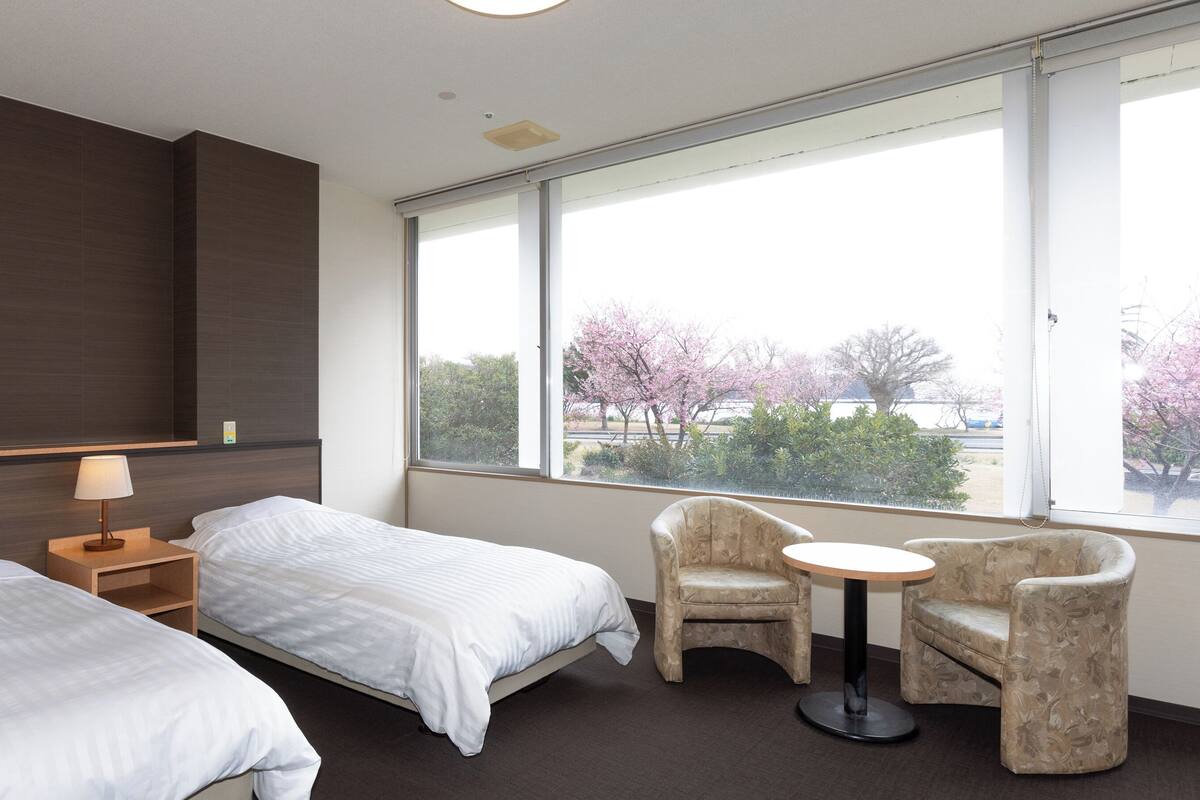 Ein Hotelzimmer mit zwei Betten, einem kleinen Tisch, einem Stuhl und einem großen Fenster mit Blick auf Bäume.