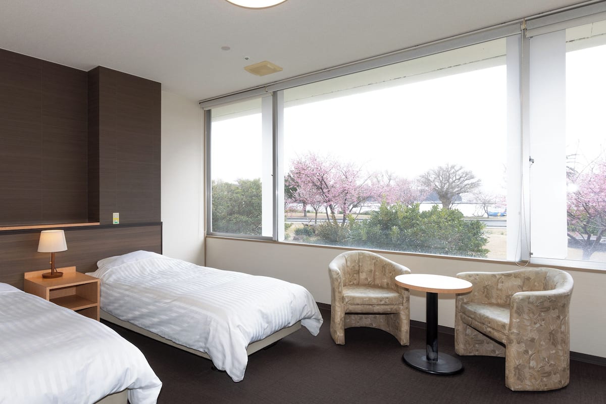 Quarto de hotel com duas camas, uma mesa pequena, uma cadeira e uma janela grande com vista para árvores.