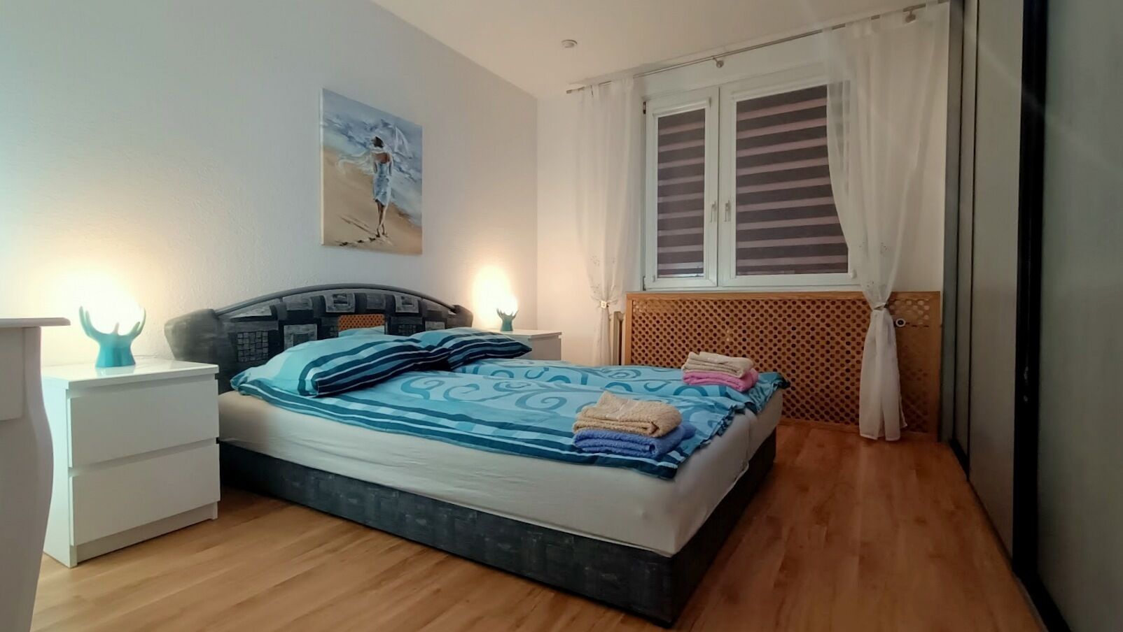 Un dormitorio con cama, mesitas de noche, un cuadro en la pared y una ventana con cortinas.