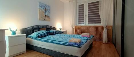 Un dormitorio con cama, mesitas de noche, un cuadro en la pared y una ventana con cortinas.