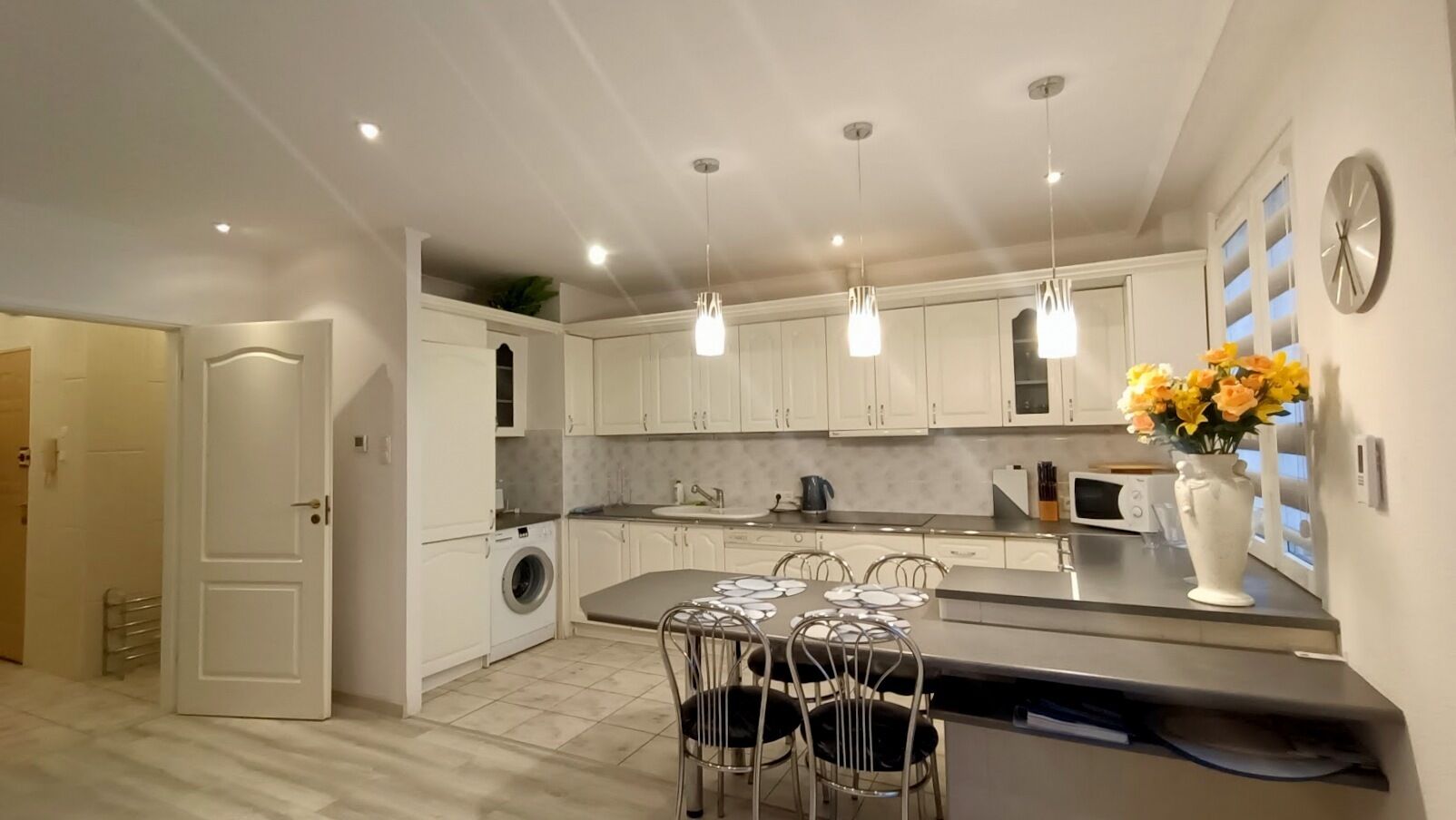 A modern kitchen with white cabinets, a built-in washing machine, a dining area with a table and chairs, pendant lights, and a vase with flowers on the counter.