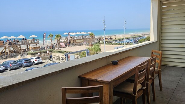 Een balkon met een houten tafel en stoelen, met uitzicht op een strand met parasols en auto's.