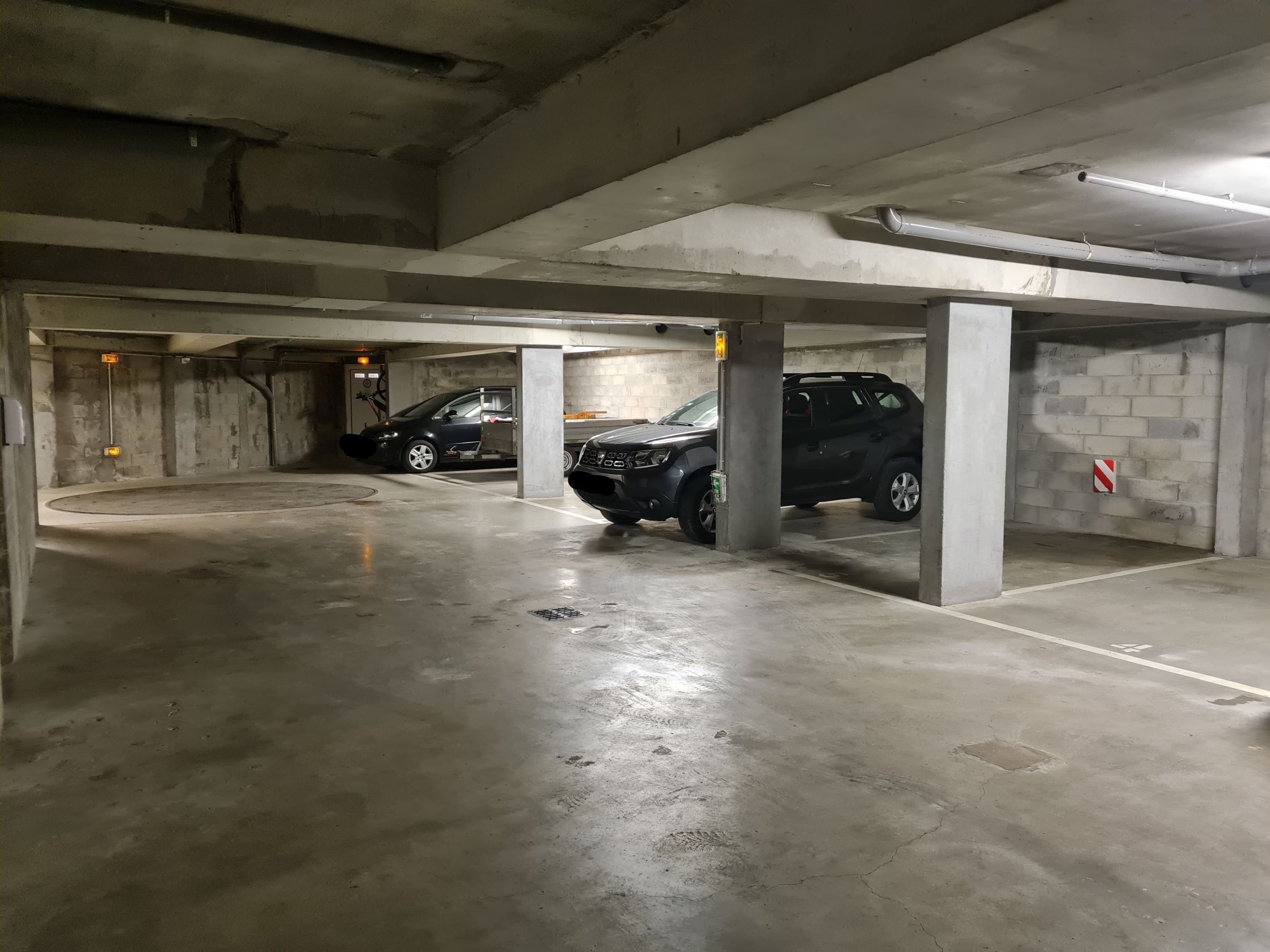 A multi-level parking garage with concrete floors and walls, featuring several parked cars and visible structural elements like columns and ceiling pipes.
