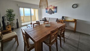 Een ruime woonkamer met een houten eettafel, stoelen, een bank en een salontafel. Er is een groot raam met uitzicht op de omgeving.
