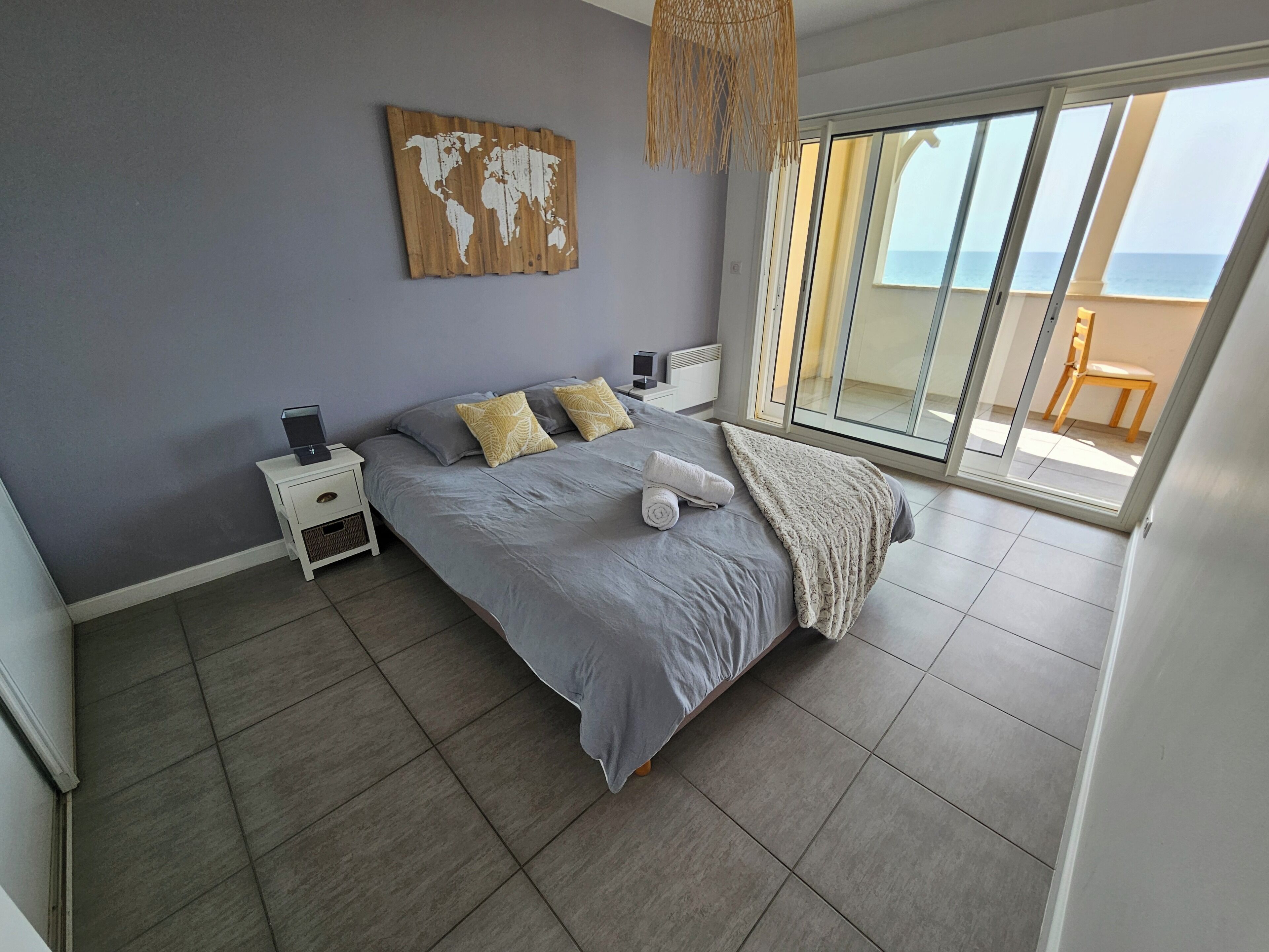 A bedroom with a large bed, a world map on the wall, a nightstand, and a view of the sea through a sliding door.