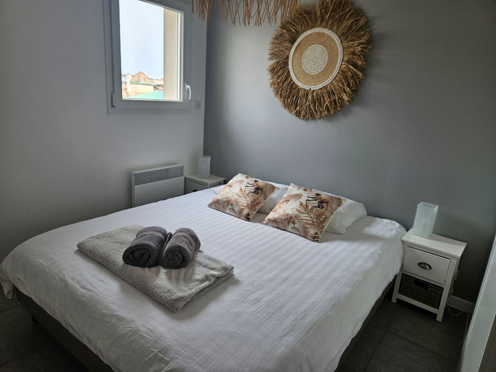 A neatly made bed with rolled towels, a decorative wall hanging, and a bedside table with a tissue box.