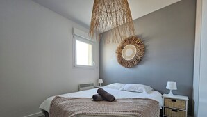 A bedroom with a large bed, a bedside table with a lamp, a wall-mounted circular woven decoration, and a window with natural light. - Pasino bright apartment,+Terrace15m²,face mer, WIFI, Parking included, 8p,N°6 (Lacanau)