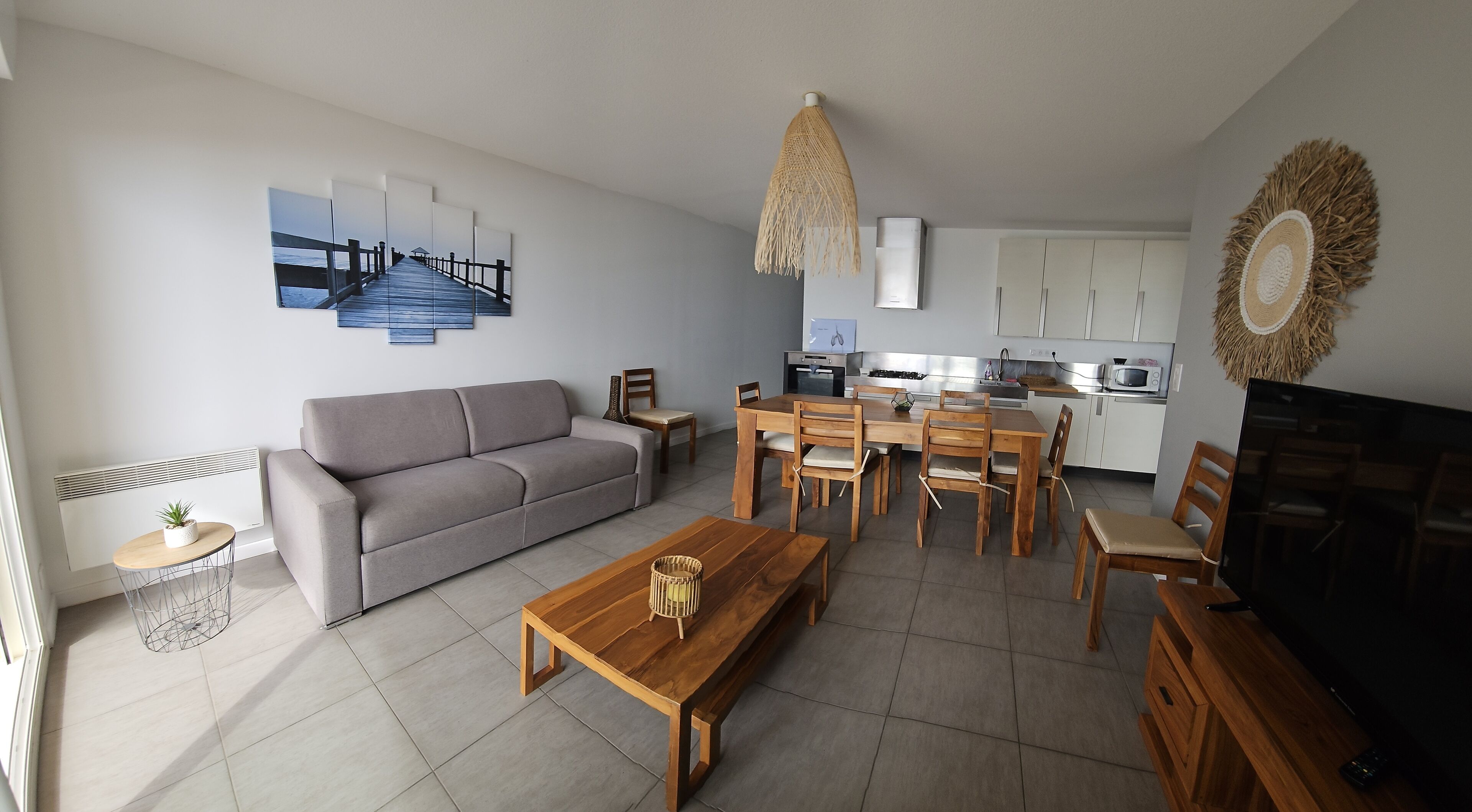 Ein modernes Wohnzimmer mit grauem Sofa, einem Holzespeisetisch mit Stühlen, einem Fernseher und einer Küche im Hintergrund.