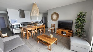 A modern living room with a wooden dining table, grey sofas, a TV, and a kitchen in the background. - Pasino bright apartment,+Terrace15m²,face mer, WIFI, Parking included, 8p,N°6 (Lacanau)