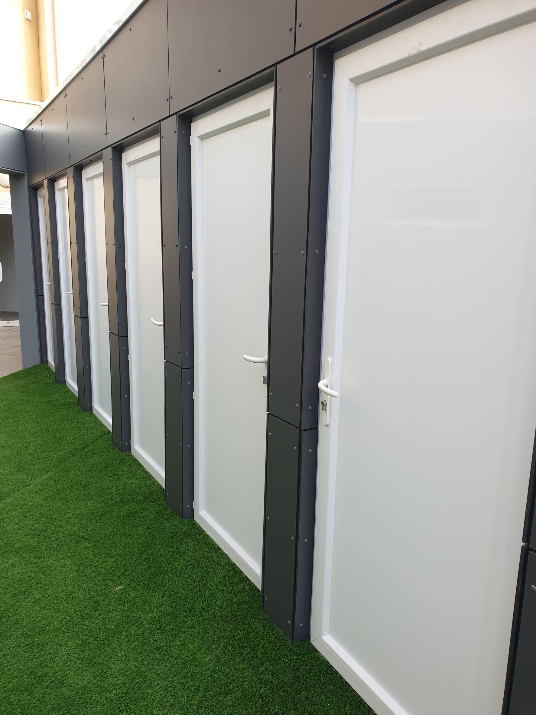 A row of closed doors with a uniform design, set against a wall with a green artificial turf in front.