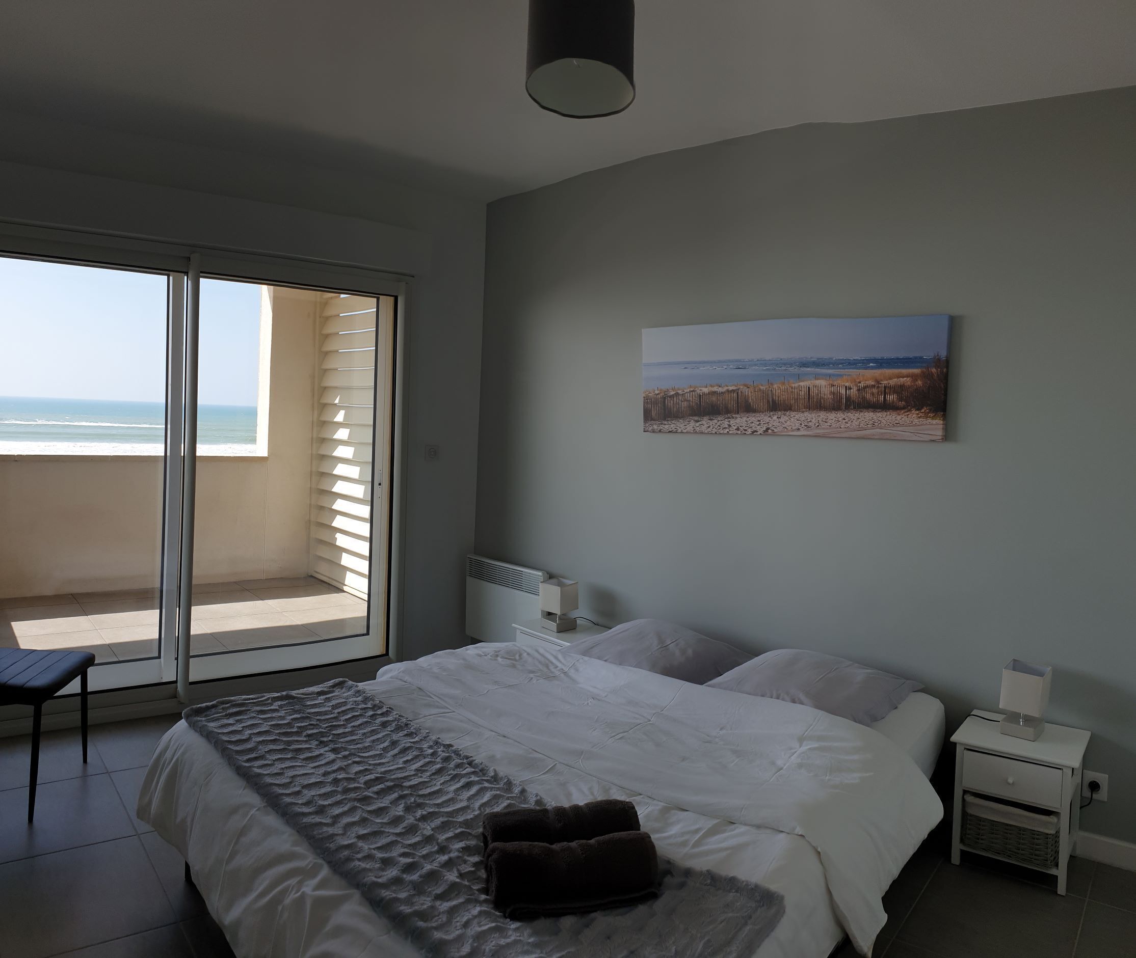 Quarto com uma cama grande, vista para a praia através de uma porta de correr, mesa de cabeceira com abajur e um quadro na parede.
