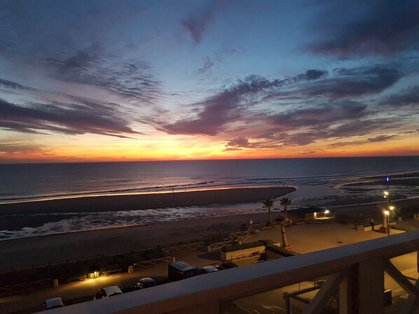 Vista del atardecer desde un balcón con vista a una playa, un estacionamiento y sillas de descanso.