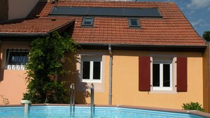 A swimming pool with a tiled deck, a house with a red-tiled roof, and a window with red shutters. - Between Colmar and Grand Ballon 
Townhouse with garden and double garage (Guebwiller)