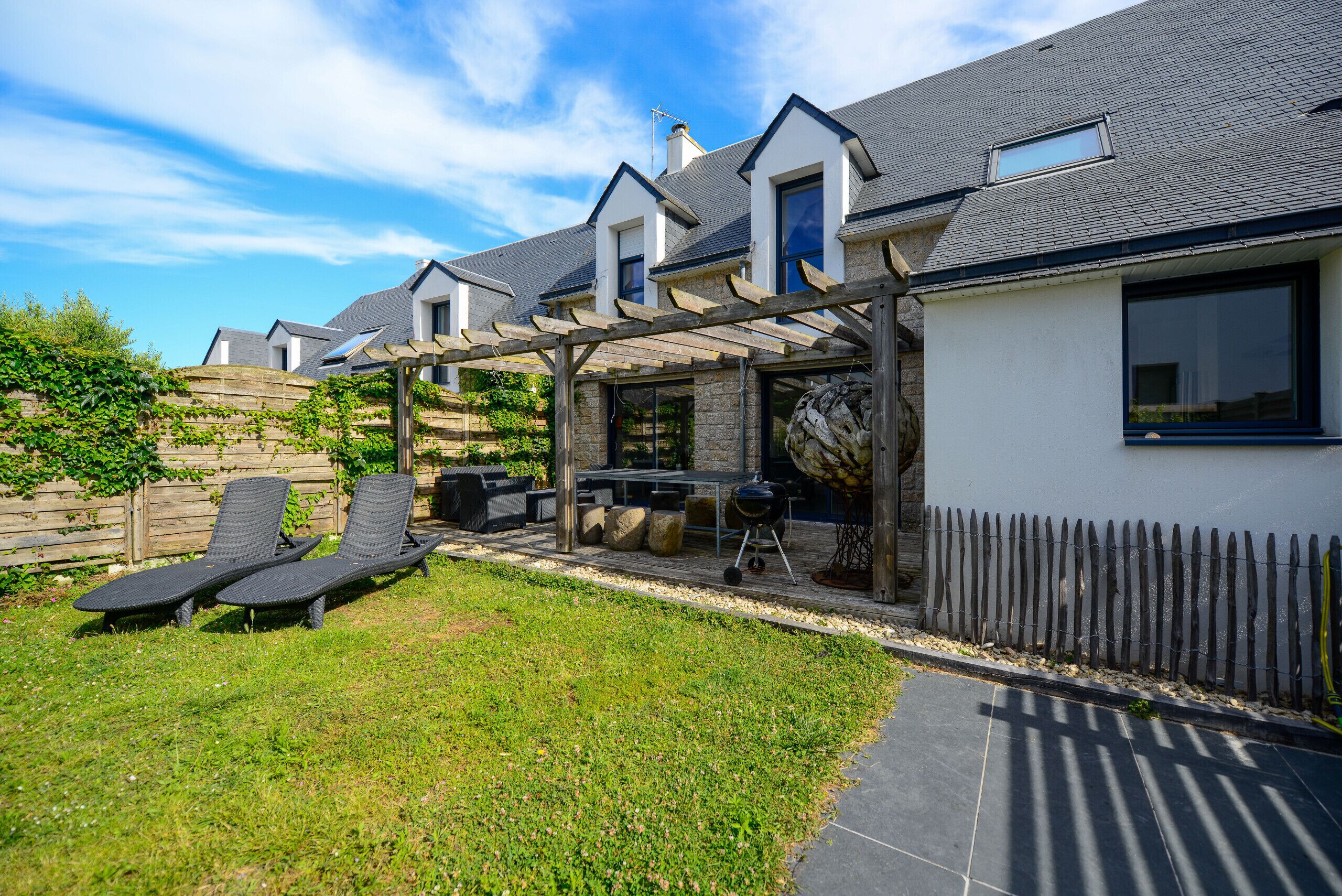 Una casa moderna con un patio que incluye dos sillones, una parrilla y una pérgola.
