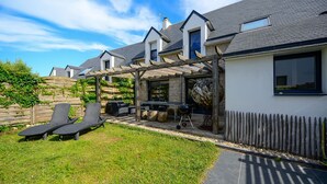 A modern house with a patio area featuring two lounge chairs, a barbecue grill, and a pergola.
