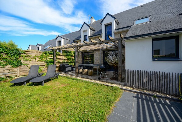 A modern house with a patio area featuring two lounge chairs, a barbecue grill, and a pergola.
