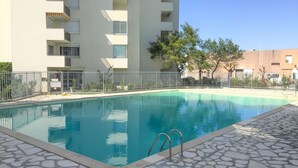 A swimming pool with a stone-tiled deck, surrounded by a metal fence and a multi-story building with balconies.