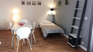 A compact room with a dining table and chairs, a bed, a bedside lamp, and a shelf unit.