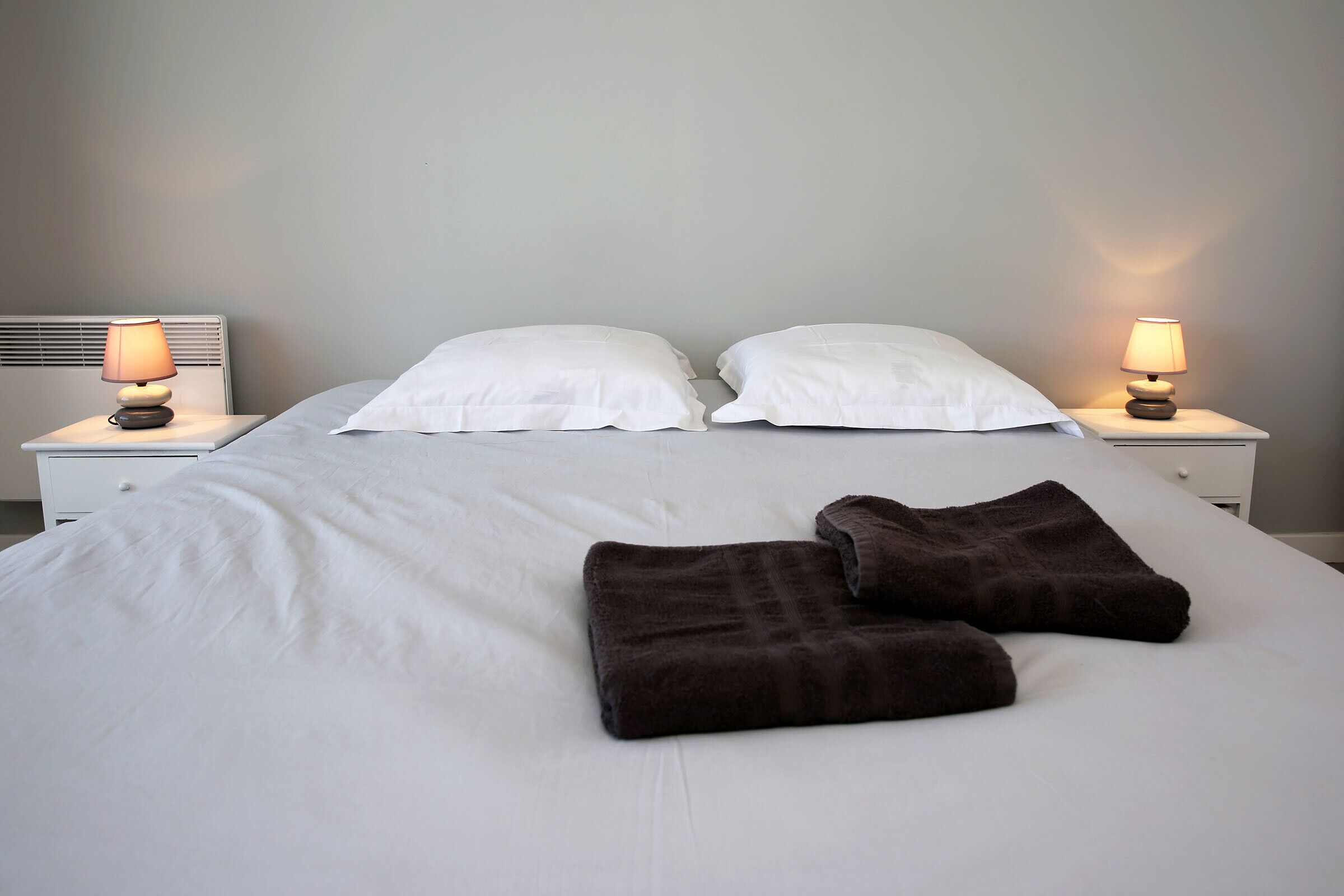 A neatly made bed with white sheets and two brown pillows, flanked by two bedside lamps on nightstands.