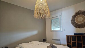 A bedroom with a bed, a bedside table with a lamp, a wall-mounted heater, a window with blinds, and a decorative wall hanging. - Pasino bright apartment +Terrace15m2, seafront, WIFI, Parking included, 6p,N°5 (Lacanau Océan)