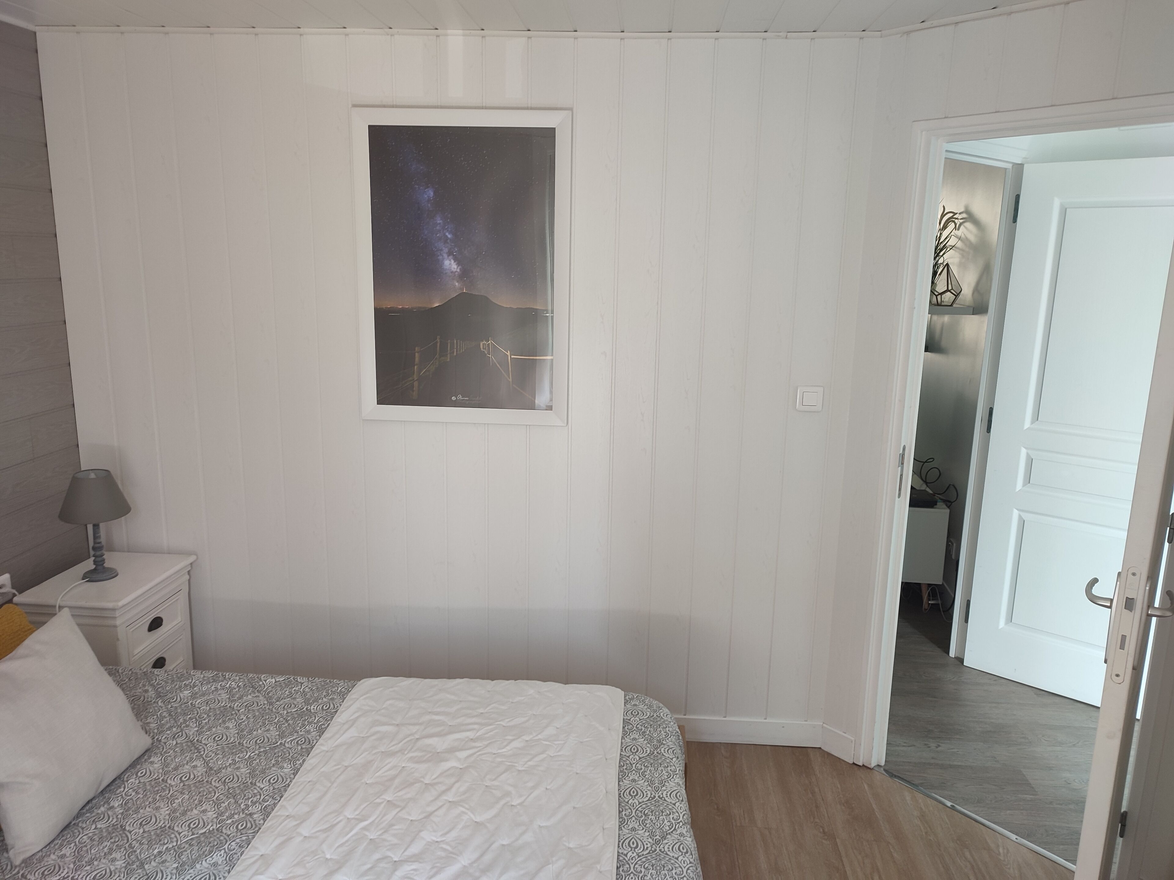 Ein Schlafzimmer mit einem Bett, einem Nachttisch, einer Lampe, einem Bilderrahmen mit einem Nachthimmel und einer offenen Tür, die zu einem Flur führt.