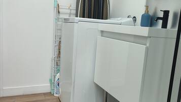 A compact laundry room with a washing machine, drying rack, and a small shelf with toiletries.