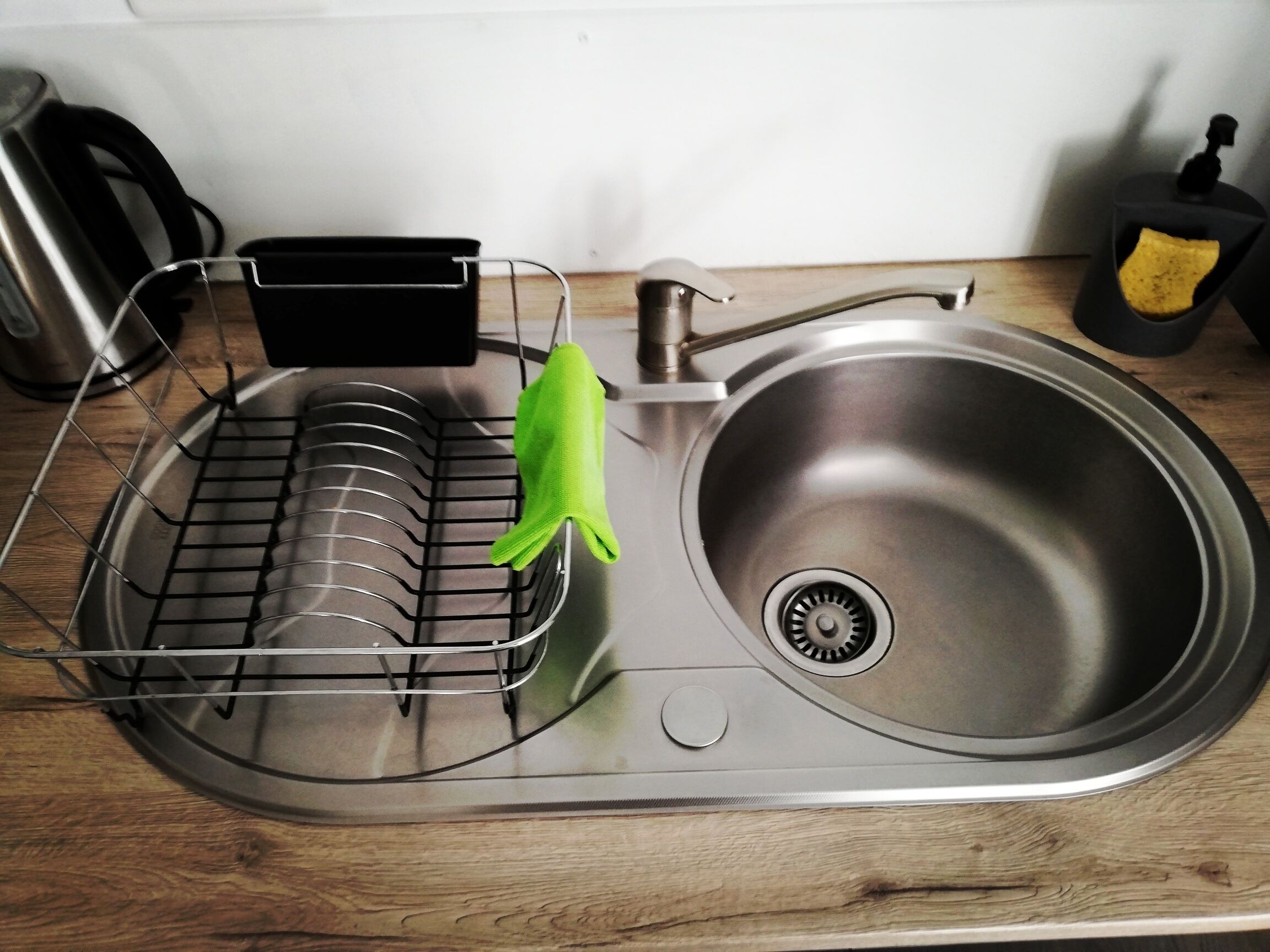 A stainless steel kitchen sink with a dish drying rack and a green cloth hanging on a rack.