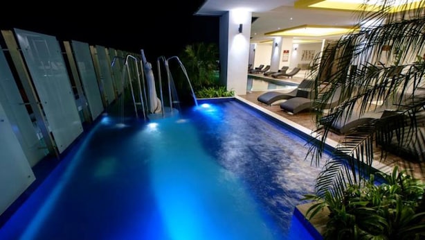 Une piscine intérieure dont les bords sont illuminés en bleu, entourée de chaises longues et de plantes tropicales.