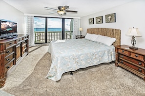 A bedroom with a bed, a television, a ceiling fan, a nightstand, a lamp, and a view of the ocean through a sliding door.