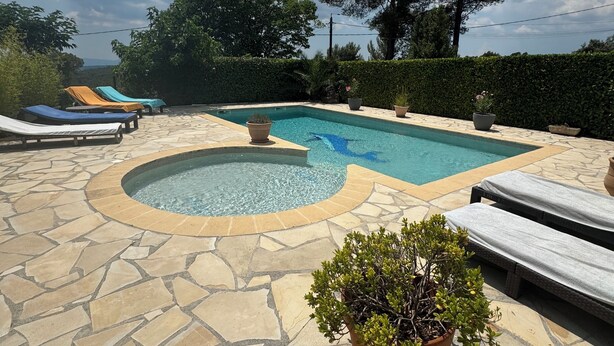 A swimming pool with lounge chairs and a hot tub, surrounded by stone tiles and greenery.