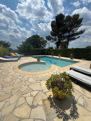 A swimming pool with lounge chairs and a hot tub, surrounded by stone tiles and greenery.
