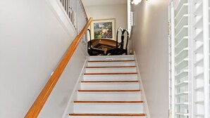 Un escalier avec des marches blanches et des rampes en bois, menant à un palier où se trouvent une chaise et un luminaire fixé au mur.