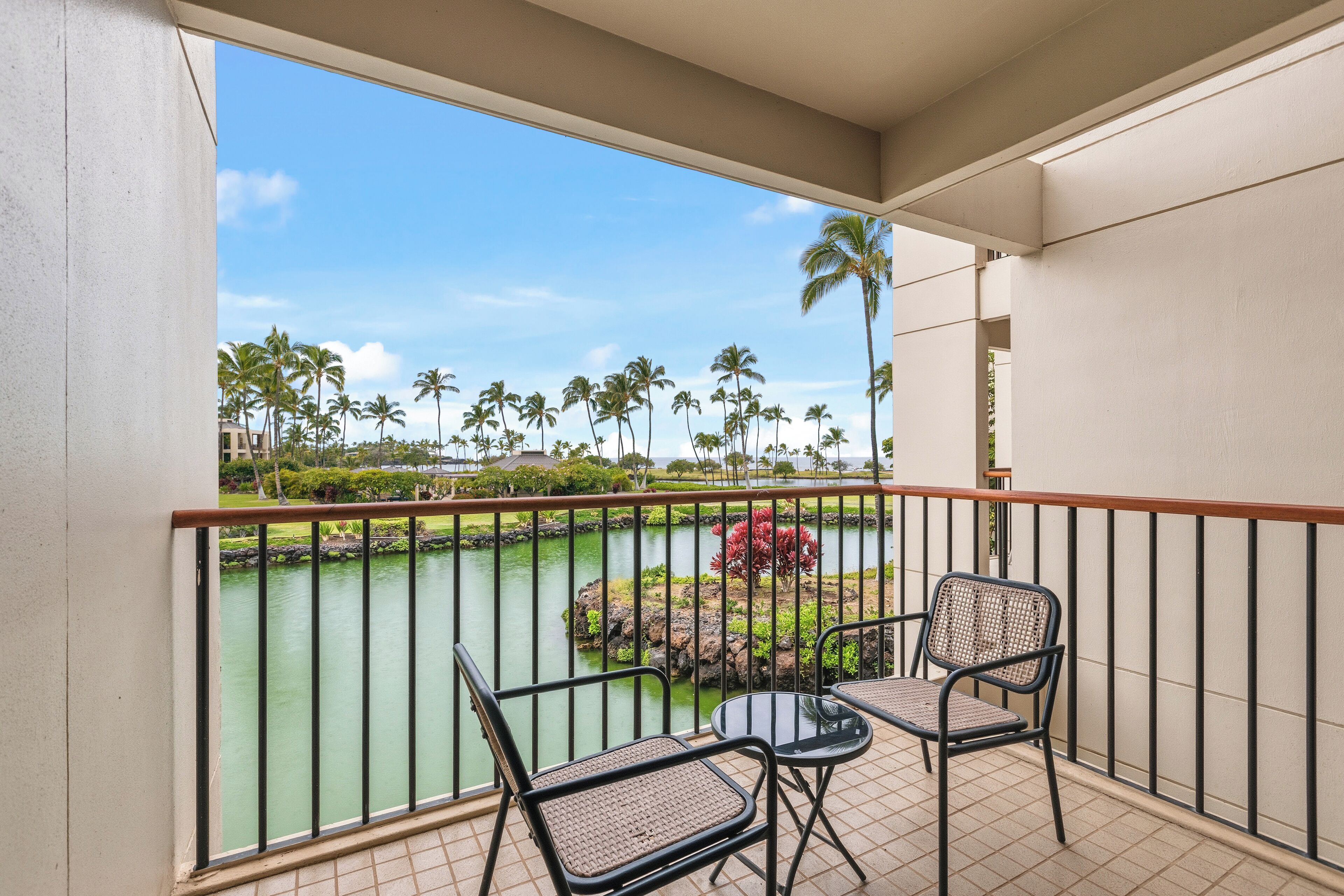A balcony with two wicker chairs, a small table, and a view of a water body with palm trees and a red bush.
