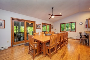 A dining area with a wooden table and chairs, a ceiling fan, and large windows with a view of greenery.