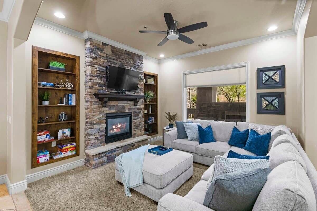 A living room with a stone fireplace, a flat-screen TV, a ceiling fan, and a large gray sectional sofa with blue cushions.