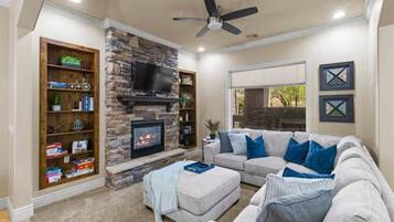 A living room with a stone fireplace, a flat-screen TV, a ceiling fan, and a large gray sectional sofa with blue cushions.