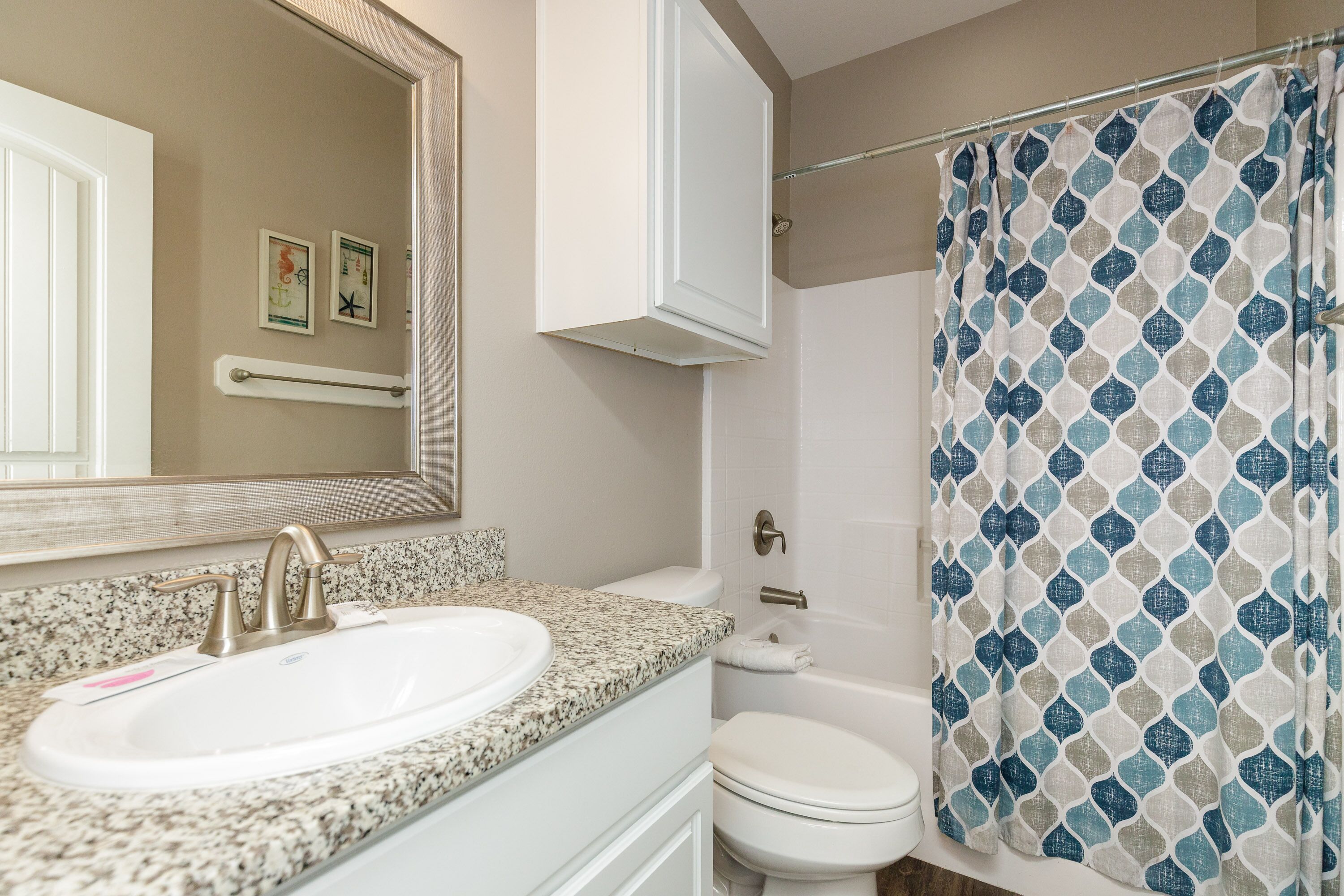Une salle de bain avec un lavabo sur un plan de travail en granit, des toilettes blanches, un rideau de douche à motifs géométriques et un miroir au-dessus du lavabo.