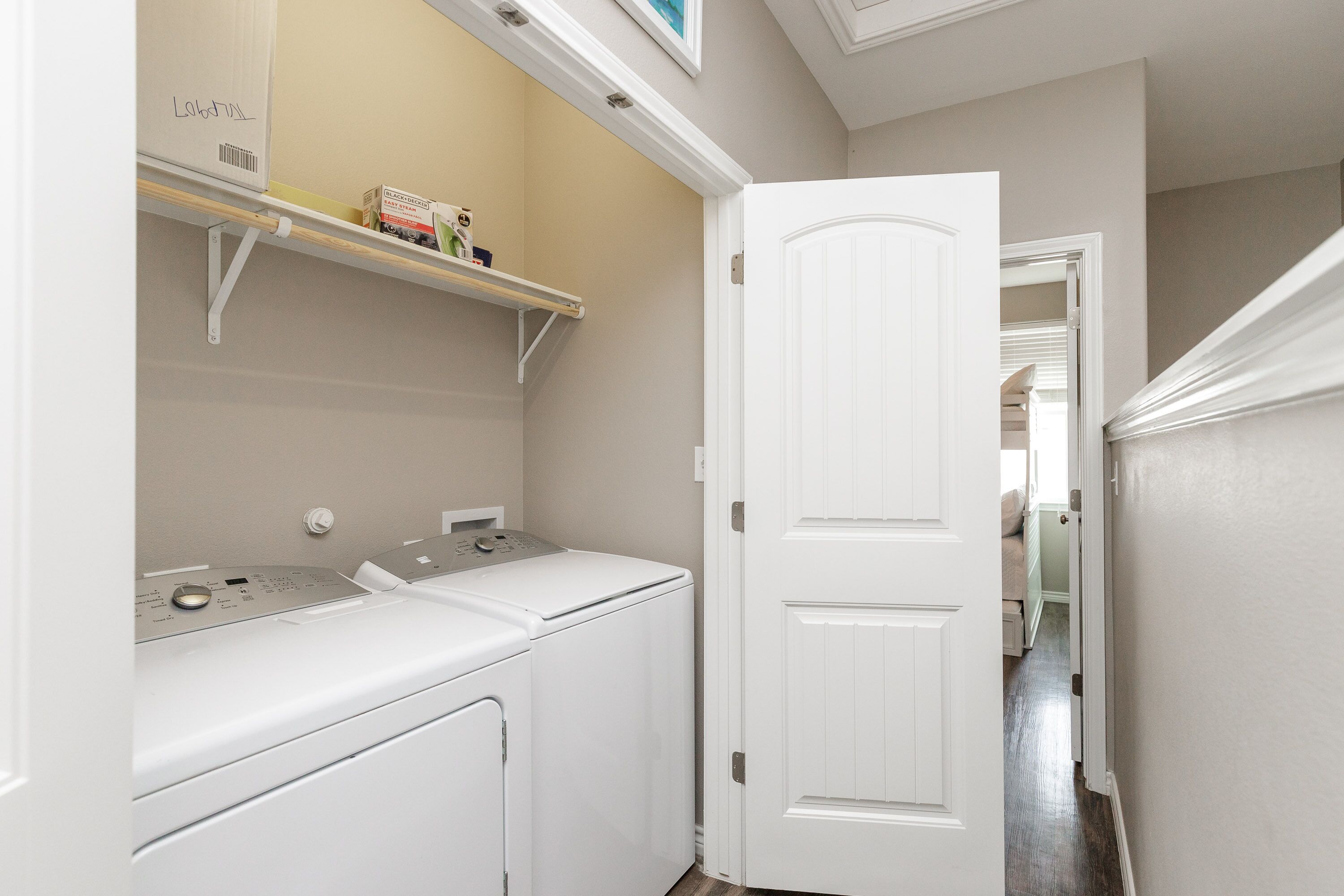 A laundry room with a washing machine and dryer, a shelf with toiletries, and a door leading to another room.