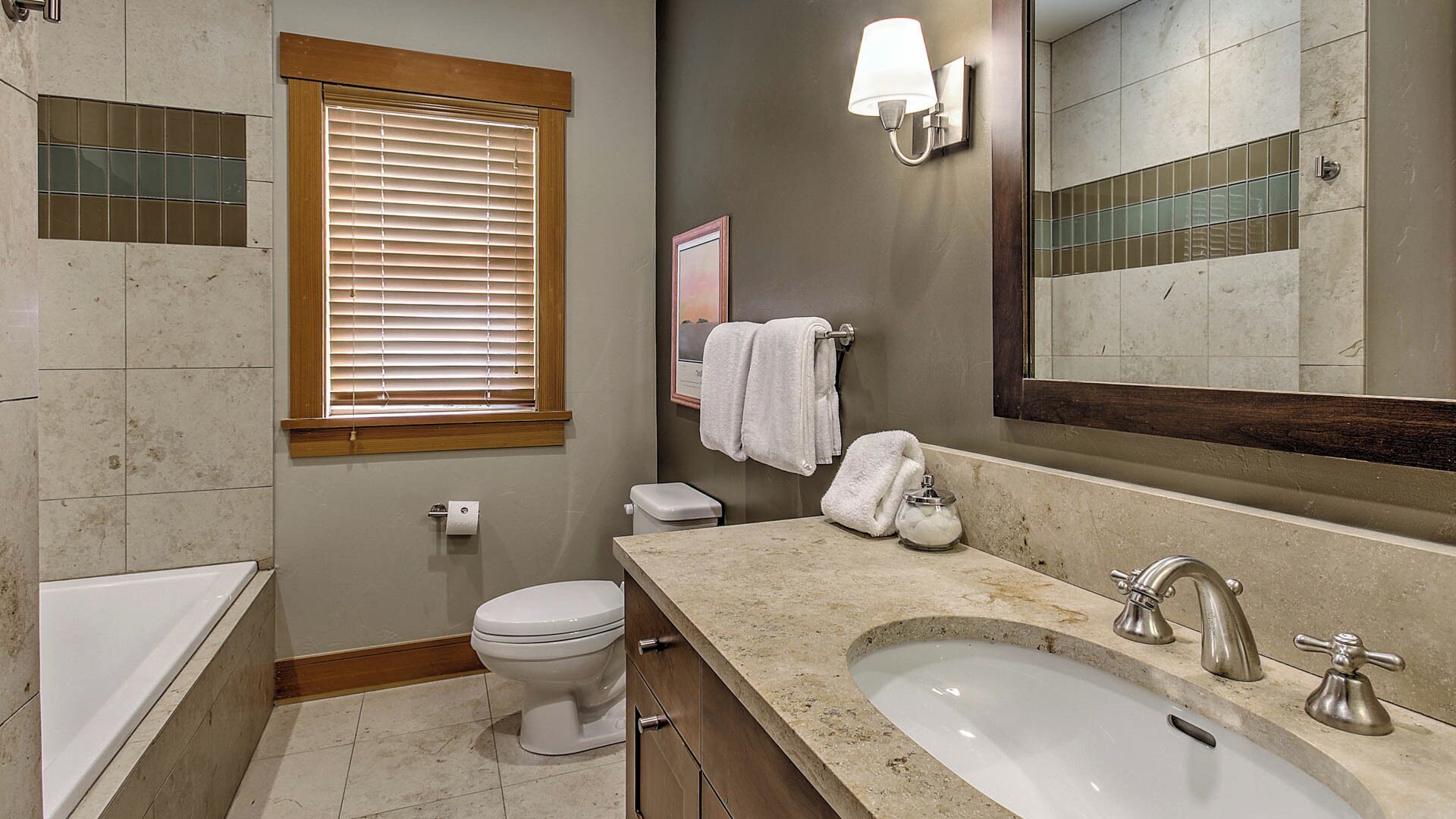A bathroom with a toilet, sink, and bathtub, featuring a window with blinds, a towel rack, and a mirror.