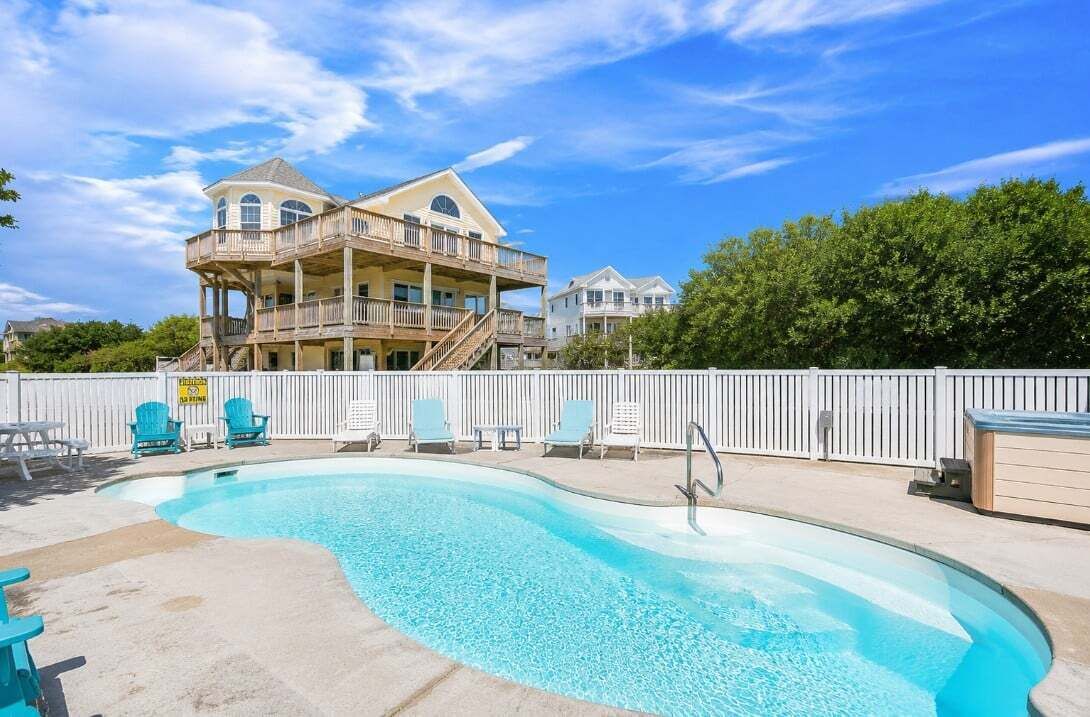 A swimming pool with lounge chairs, a multi-level building with balconies, and a white fence.
