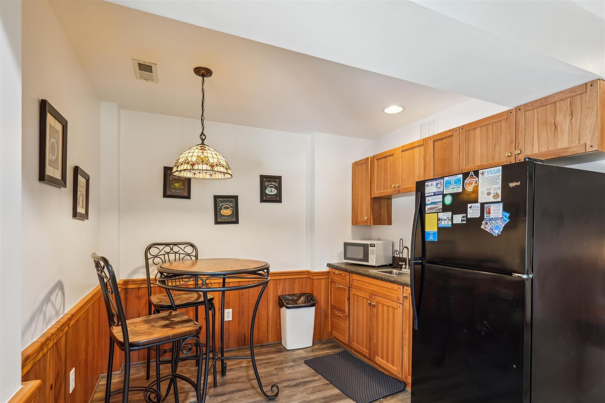A kitchen with wooden cabinets, a black refrigerator, a microwave, a dining table with chairs, and a hanging light fixture.