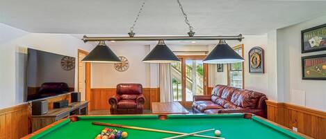 Ein Billardtisch mit Kugeln, ein Fernseher und zwei Ledersessel in einem Raum mit Holzwänden und einer Wand mit Billard-Motiven.
