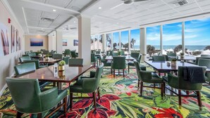 A conference room with tables and chairs, large windows with a view of the beach, and a ceiling fan.