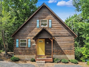 Une cabane en bois avec une porte jaune, deux fenêtres avec des volets et un petit porche.