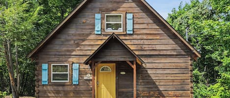 Una cabaña de madera con puerta amarilla, dos ventanas con contraventanas y un pequeño porche.