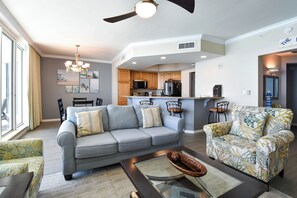 A modern living room with a grey sofa, a floral armchair, and a glass coffee table. A kitchen with wooden cabinets and a microwave is visible in the background. - A three-bedroom beachfront, modern Gulf Coast getaway! 🌴 (Destin)
